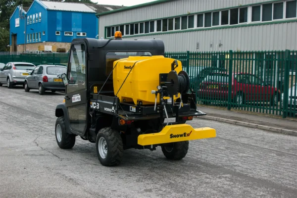 Wessex snowex liquid de-icing system | vss-1000 is a small utility vehicle featuring a yellow spreader attachment, is parked on a paved road near a fenced industrial building. Several parked cars are visible in the background.