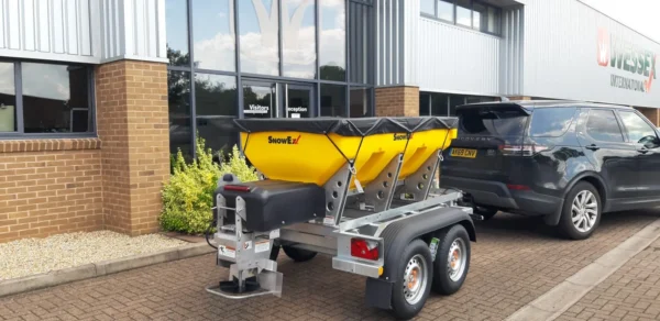 Wessex Snowex Helixx Spreaders | Middlewich Machinery A yellow wessex snowex helixx spreader on a dual-axle trailer is hitched to a black suv parked outside a commercial building.