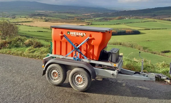 A wessex polar one salt and grit spreader, painted in orange, is parked on the roadside with a vast, green hilly landscape in the background.
